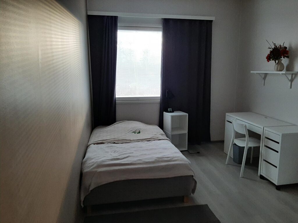 Sisustettu huone. Iso ikkuna, sänky, pöytä ja tuoli.