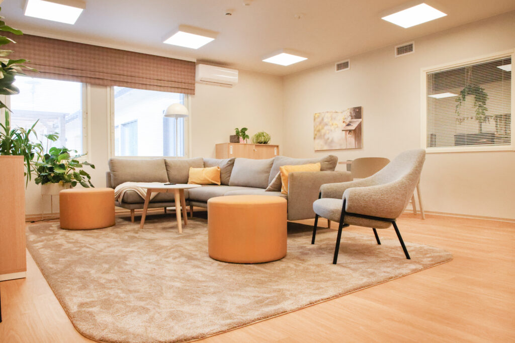 Oleskelutila, jossa harmaa kulmasohva, kasveja, iso vaalea matto.