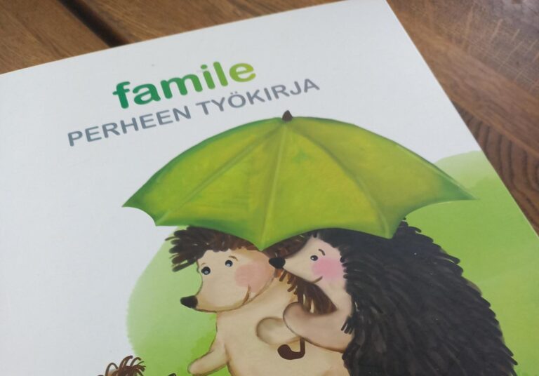 Famile perheen työkirja puupöydällä. Valkoinen kirja, jossa piirretyt siilit kannessa pitelevät vihreää sateenvarjoa ja pehmopupua.